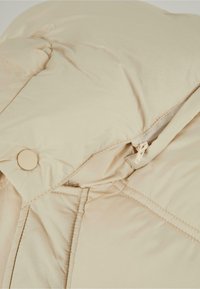 Beige puffer jacket with a smooth, padded texture. Features a zip closure and button accents near the collar. Quilted design visible.
