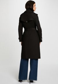 Donna con capelli scuri che indossa un trench coat nero con cintura, jeans larghi blu e scarpe con tacco nere, rivolta di spalle su uno sfondo semplice.