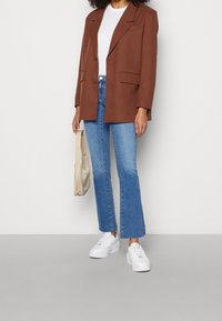 Mujer con blazer marrón, blusa blanca, jeans de campana cortos azules, zapatillas blancas y sosteniendo un pequeño bolso beige a su lado.