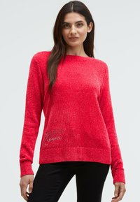 Frau in einem leuchtend roten, strukturierten Langarmpullover mit schwarzer Hose, die vor einem einfachen, hellen Hintergrund steht.