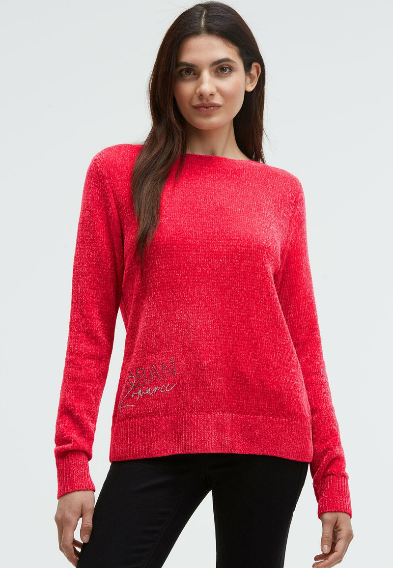 Frau in einem leuchtend roten, strukturierten Langarmpullover mit schwarzer Hose, die vor einem einfachen, hellen Hintergrund steht.