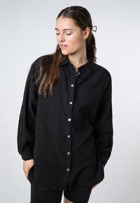 Black button-up shirt made from textured fabric. Features a collar, long sleeves, and blue buttons. Loose fit and curved hem.