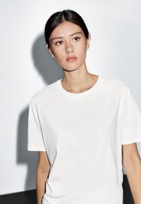 Bílý bavlněný tričko s kulatým výstřihem a krátkými rukávy, vystavené na minimalistickém pozadí, vrhající měkký stín.