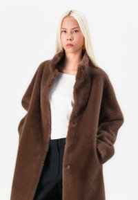 Manteau en fausse fourrure marron avec un col, deux poches avant et une coupe décontractée. Porté sur un haut blanc et un pantalon noir, avec une texture douce.