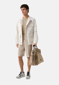 Veste utilitaire légère en crème avec plusieurs poches, pull beige, short beige et baskets grises. Tenant un grand sac de voyage beige.