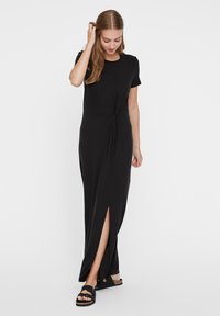 Μαύρο maxi φόρεμα με κοντά μανίκια, λεπτομέρεια με κόμπους στο μπροστινό μέρος, πλαϊνή σχισμή και μαλακό ύφασμα από μείγμα βαμβακιού. Φορεμένο με μαύρα σανδάλια.