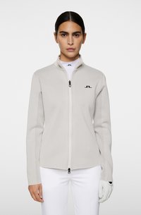 Veste zippée gris clair avec un col haut, présentant un tissu texturé et des accents en mesh sur les manches. Logo noir sur la poitrine gauche.
