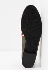 Semelle de chaussure avec un fond en caoutchouc noir texturé, tige en daim vert olive et une bande décorative en rouge, blanc et noir.