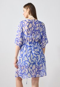 Vestido de cobertura translúcido con patrones abstractos en azul y beige, mangas cortas, cintura elástica y lazos ajustables en el cuello.