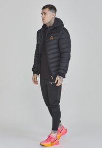 Doudoune noire avec capuche et logo brodé, associée à un jogging noir slim-fit et des chaussures de course roses vives.