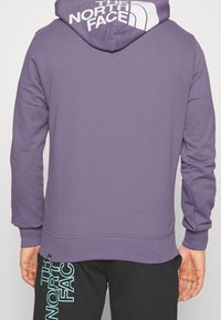 Lila hoodie med vit logga på ryggen, med dragsko i huvan och ribbade manschetter. Parat med svarta joggers som visar logga i teal.