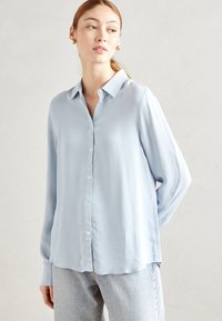 Donna con camicia button-down a maniche lunghe blu chiaro e jeans chiari che guarda di lato su uno sfondo neutro.