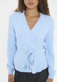 Sieviete valkā gaiši zilo adījuma jaciņu ar V-veida kaklu un lentu, apvienojot to ar melnu svārku.