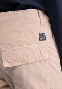 Beżowe spodnie cargo z kieszenią z klapą i czarną naszywką logo Alpha Industries, noszone z ciemnogranatową górą.