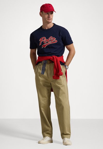 Jūras kara flotes t-kreklis ar sarkano "Polo" logo, khaki bikses, sarkans džemperis ap vidukli, sarkans cepure un baltas kedas. Ikdienišķs apģērbs ar klasiskām sastāvdaļām.