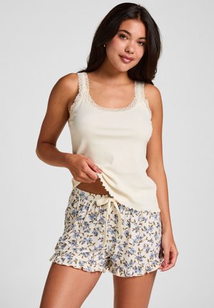 Jonge vrouw draagt een crèmekleurig tanktop met kanten rand en korte broek met bloemenprint en trekkoord, staand tegen een effen lichte achtergrond.