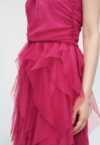 Gros plan sur une robe en tulle rose à volants montrant les détails des couches de tissu et une main reposant à côté de la jupe sur un fond uni.