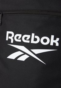 Črna platnena torba z izstopajočim belim logotipom Reebok in modernim dizajnom, z zadrgo in teksturirano površino.