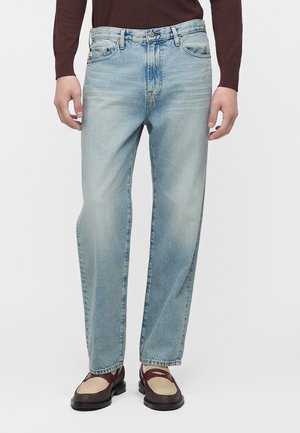 Homme portant un jean droit bleu clair, une chemise marron à manches longues et des mocassins marron foncé avec un détail en tissu beige.