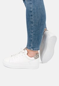 Zapatillas deportivas blancas con material texturizado, con un detalle de estampado de leopardo en el talón. Suela gruesa y diseño de punta redonda, combinadas con jeans azules.