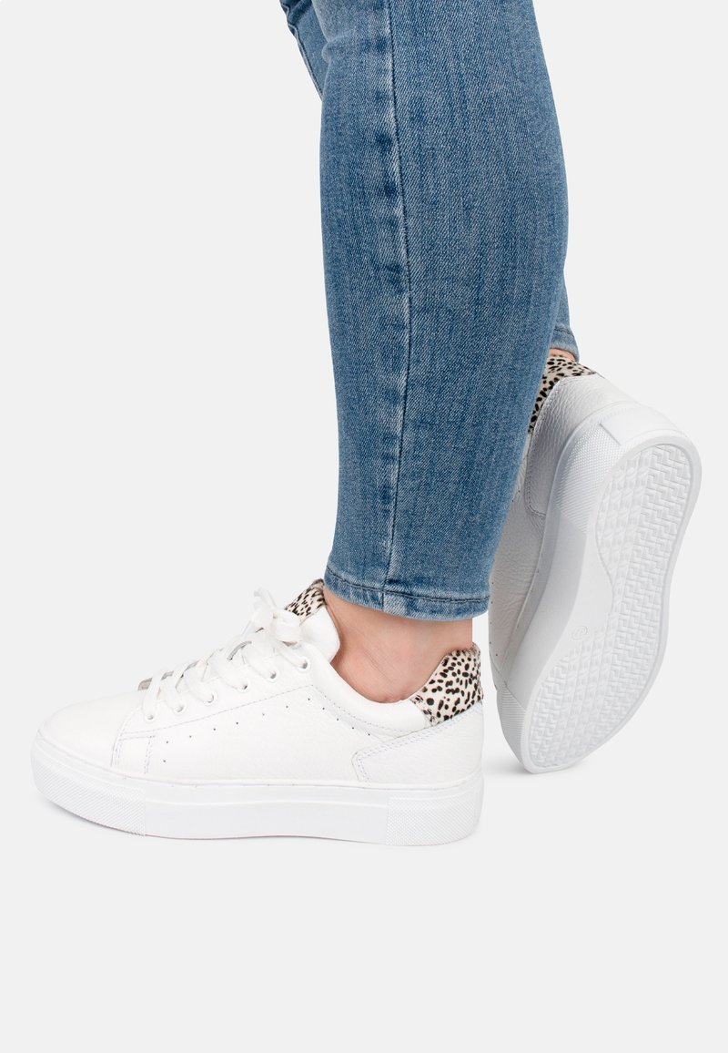 Zapatillas deportivas blancas con material texturizado, con un detalle de estampado de leopardo en el talón. Suela gruesa y diseño de punta redonda, combinadas con jeans azules.