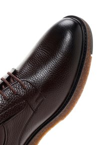 Gros plan sur une chaussure de ville en cuir marron foncé avec une surface texturée, des lacets marron et une semelle en caoutchouc brun clair sur fond blanc.