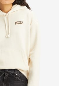 Krämmig hoodie i mjukt material med kängurufickor, snörning på huvan och en brun Levi's-logotyp på bröstet. Bärs över mörka jeans.