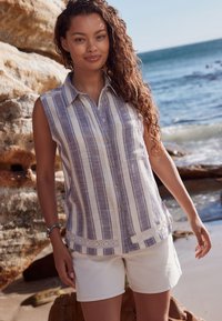 Chemisier sans manches en tissu léger à rayures verticales bleues et blanches, boutonné à l'avant, orné d'un bord en dentelle au bas. Associé à un short blanc.