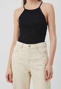 Débardeur noir à côtes avec des bretelles festonnées, associé à un jean en denim beige clair taille haute. Le tissu semble lisse et texturé.