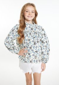 Blusa bianca con motivo floreale blu e nero, colletto arricciato e maniche a sbuffo, abbinata a pantaloni corti bianchi frangiati.