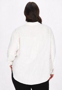 Witte lange mouwen shirt van lichtgewicht stof, met een klassieke kraag, geplooide achterkant en gebogen zoom. Zwarte broek zichtbaar aan de onderkant.
