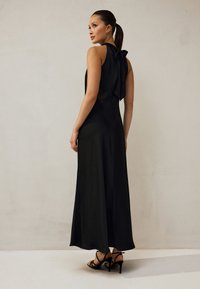 Schwarzes Maxikleid mit Neckholder und einer Schleifendetail am Rücken. Es verfügt über ein fließendes Design und einen dezenten strukturierten Stoff. Kombiniert mit Riemchenabsätzen.