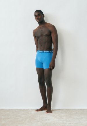 Άνδρας μοντέλο στέκεται ξυπόλητος μπροστά σε έναν λευκό τοίχο, φορώντας φωτεινά μπλε μποξεράκια Björn Borg, κοιτώντας ελαφρώς στο πλάι.