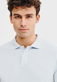 Jasnoniebieska koszulka polo z teksturowanej tkaniny, z klasycznym kołnierzykiem, trzema guzikami oraz małym haftowanym logo na piersi.