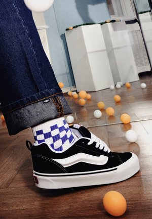 Primer plano de una persona con vaqueros con el bajo vuelto, calcetines de cuadros y zapatillas deportivas blancas y negras, entre bolas naranjas y blancas esparcidas.