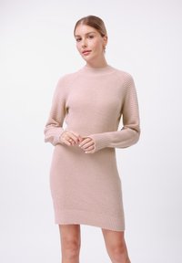 Robe pull en beige clair avec texture côtelée, col montant, manches longues et un ourlet qui descend légèrement sur les côtés.