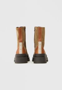 Botas de tobillo de ante marrón con un tacón marrón más oscuro, que presentan un zipper lateral y un acento de cuero en contraste. Parte superior texturizada con detalle de logotipo.