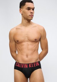 Μυώδης άνδρας που φοράει μαύρα εσώρουχα Calvin Klein με κόκκινη ζώνη μέσης με λογότυπο, στέκεται μπροστά σε απλό ανοιχτό γκρι φόντο, κοιτάζοντας προς τα δεξιά.