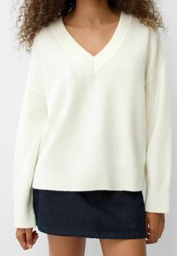 Biały sweter z dekoltem w serek, o luźnym kroju, z prążkowaną fakturą i ściągaczami. W zestawieniu z ciemną spódnicą jeansową, prezentującą swobodny fason i casualowy styl.