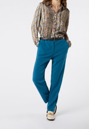 Personne portant une chemise à manches longues multicolore à motifs, rentrée dans un pantalon bleu avec une ceinture noire et des mocassins blancs avec détail chaîne dorée.