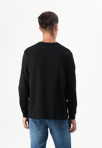 Een persoon staat met de rug naar de camera, gekleed in een zwart longsleeve shirt en blauwe spijkerbroek, tegen een effen witte achtergrond.