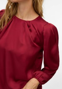 Bluse i burgunder med glatt satengfinish, rund halslinning og puffede ermer med elastiske mansjetter. Har subtile rynker ved halsen.