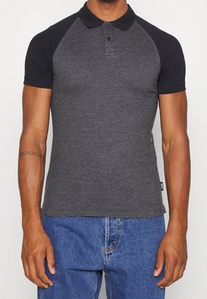 Hombre con camisa de polo de manga corta texturizada en negro y gris oscuro con cuello negro, combinado con vaqueros azules, de pie con los brazos relajados.