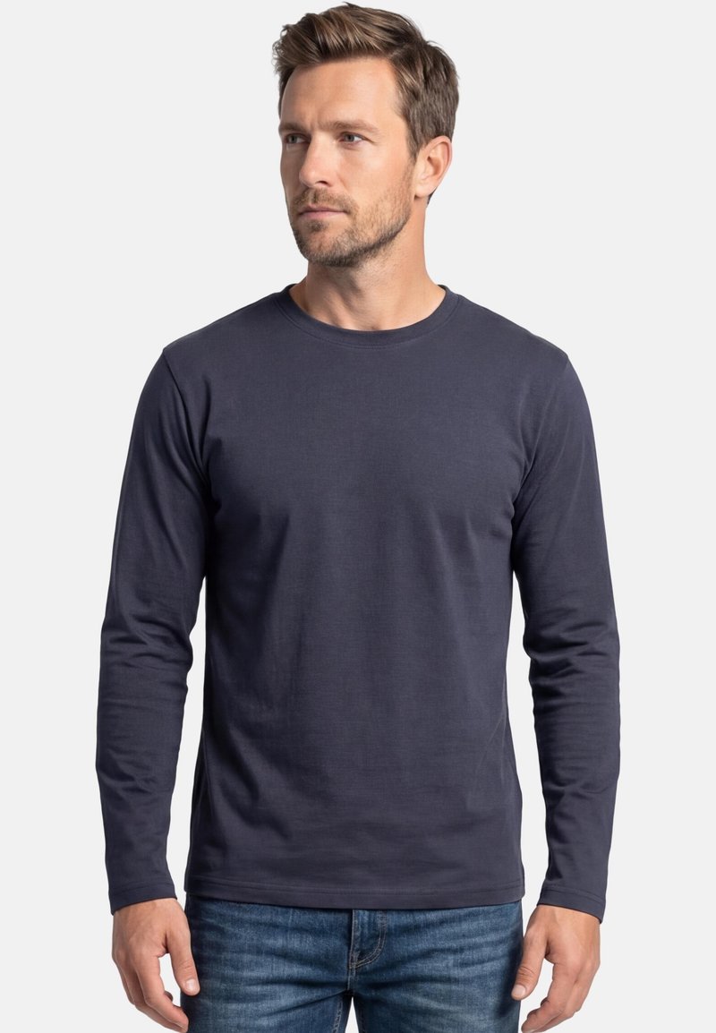 Mann mit kurzen braunen Haaren, der ein dunkelblaues Langarmshirt mit Rundhalsausschnitt und blaue Jeans trägt und vor einem schlichten weißen Hintergrund steht.