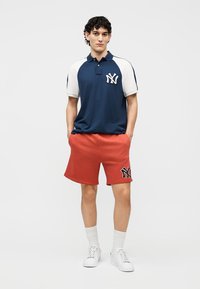 Noor noormees seisab käed taskutes, kandes tumesinist ja valget New York Yankee's polo särki ning punaseid lühkse sobiva logoga.