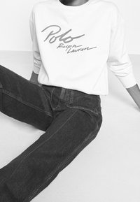 Vit sweatshirt med "Polo Ralph Lauren"-logotyp i grått bläck, kombinerad med mörka denimjeans med sömnadsdetaljer och rak passform.