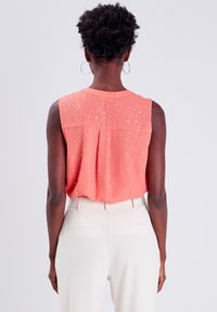 Korallenfarbene ärmellose Bluse mit subtilen Blumenmuster und lockerer Passform, kombiniert mit cremefarbenen Hosen. Die Rückansicht zeigt Designdetails.