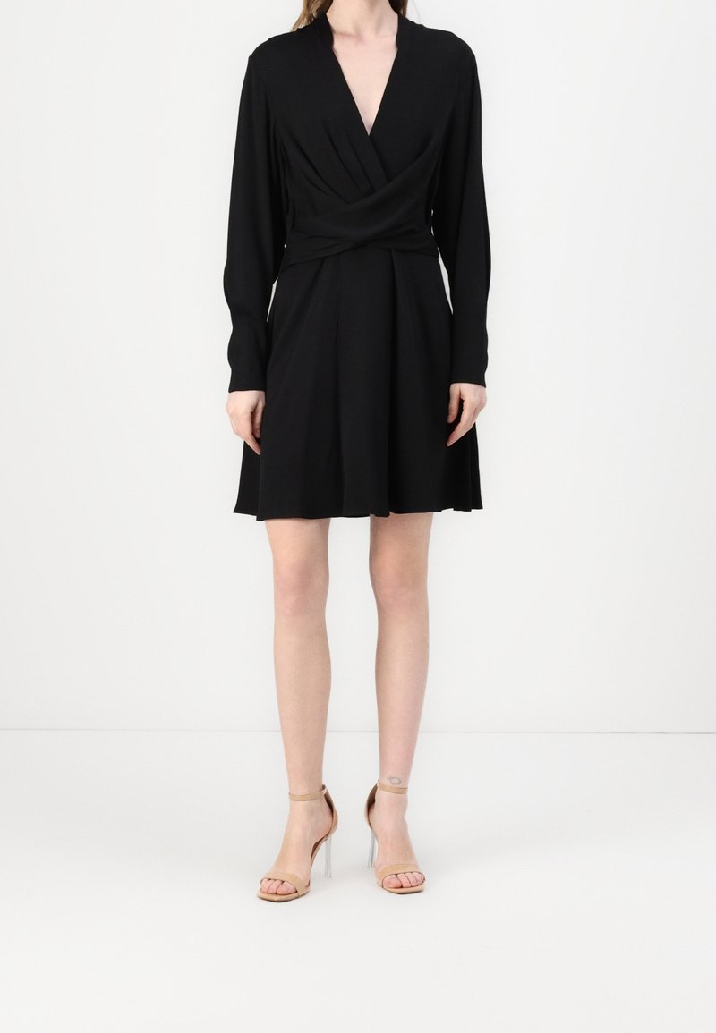 Femme portant une robe portefeuille noire manches longues avec un décolleté en V et des sandales beiges à talons hauts, debout devant un fond blanc.