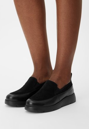 Jambes portant des chaussures noires à enfiler avec une tige en daim et des garnitures en cuir, se tenant sur une surface blanche.