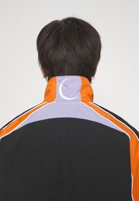 Jackpack with a high collar, featuring black fabric, orange and purple accents, white piping, and a distinctive crescent logo on the collar.